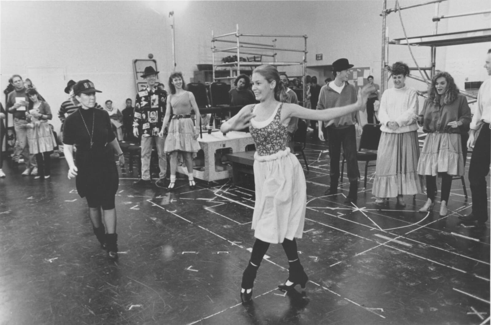A black and white photo of Susan Stroman rehearsing for Show Boat with Kim (Tammy Amerson). Other ARTISTS watch her in the background.