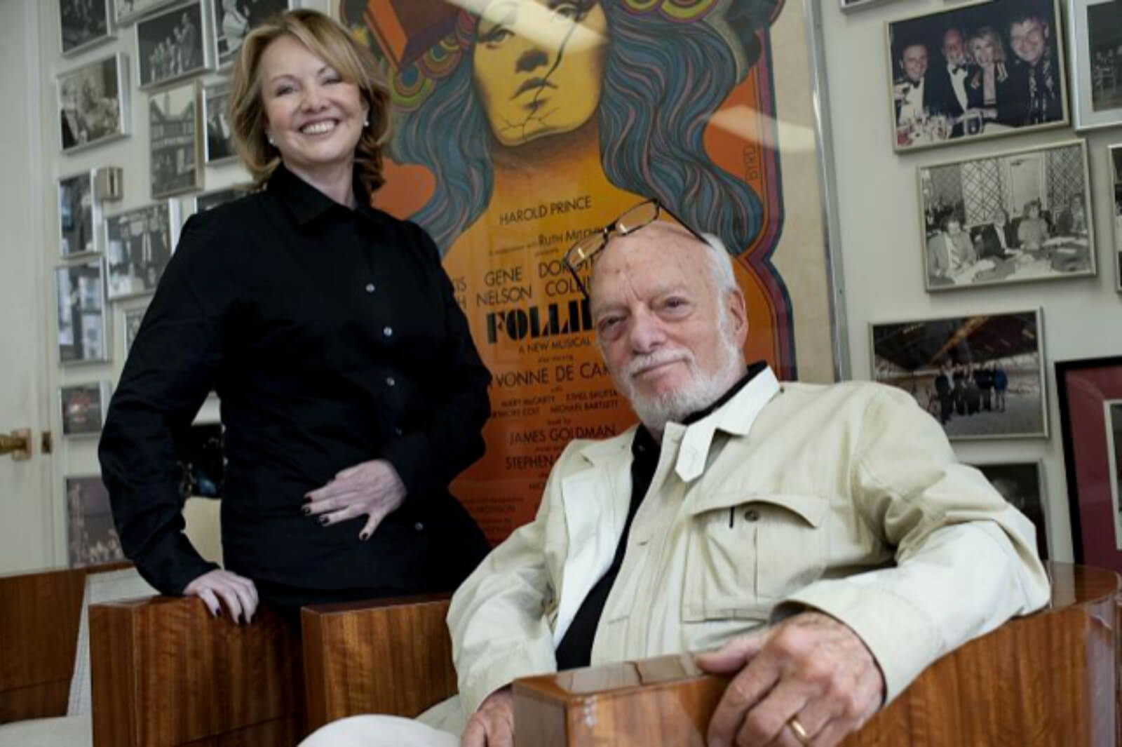 Susan Stroman in a black ensemble and Hal Prince, in a beige jacket and eyeglasses above his head, sitting, with a wall of fame in the background. They are discussing Prince of Broadway. 