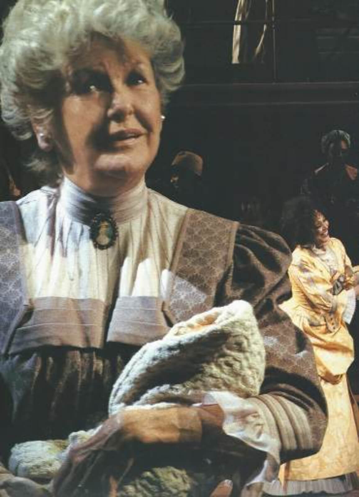 Parthy Ann Hawks (Elaine Stritch) holding a child in a blanket, and a smiling ARTIST is seen from behind her, wearing a yellow dress.