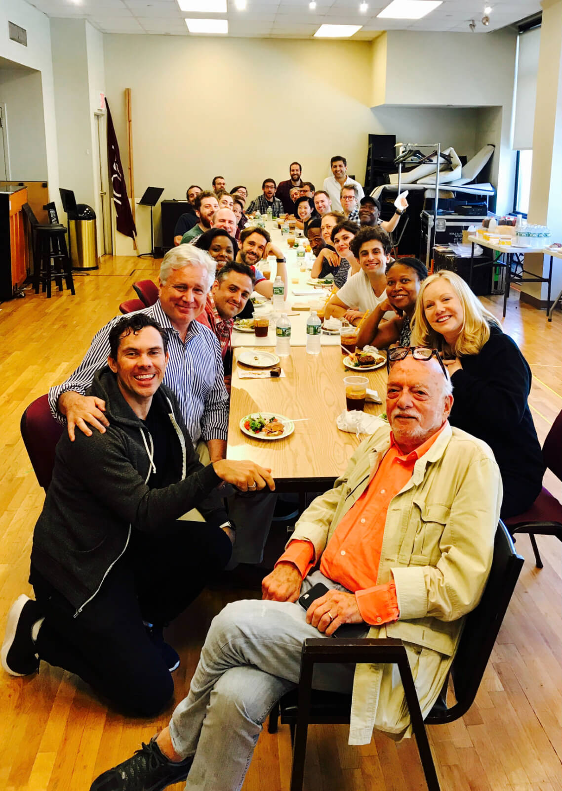 Hal Prince and Susan Stroman having lunch with the Broadway cast and creatives of Prince of Broadway. They are sitting at a long table in a rehearsal studio enjoying their lunch.
