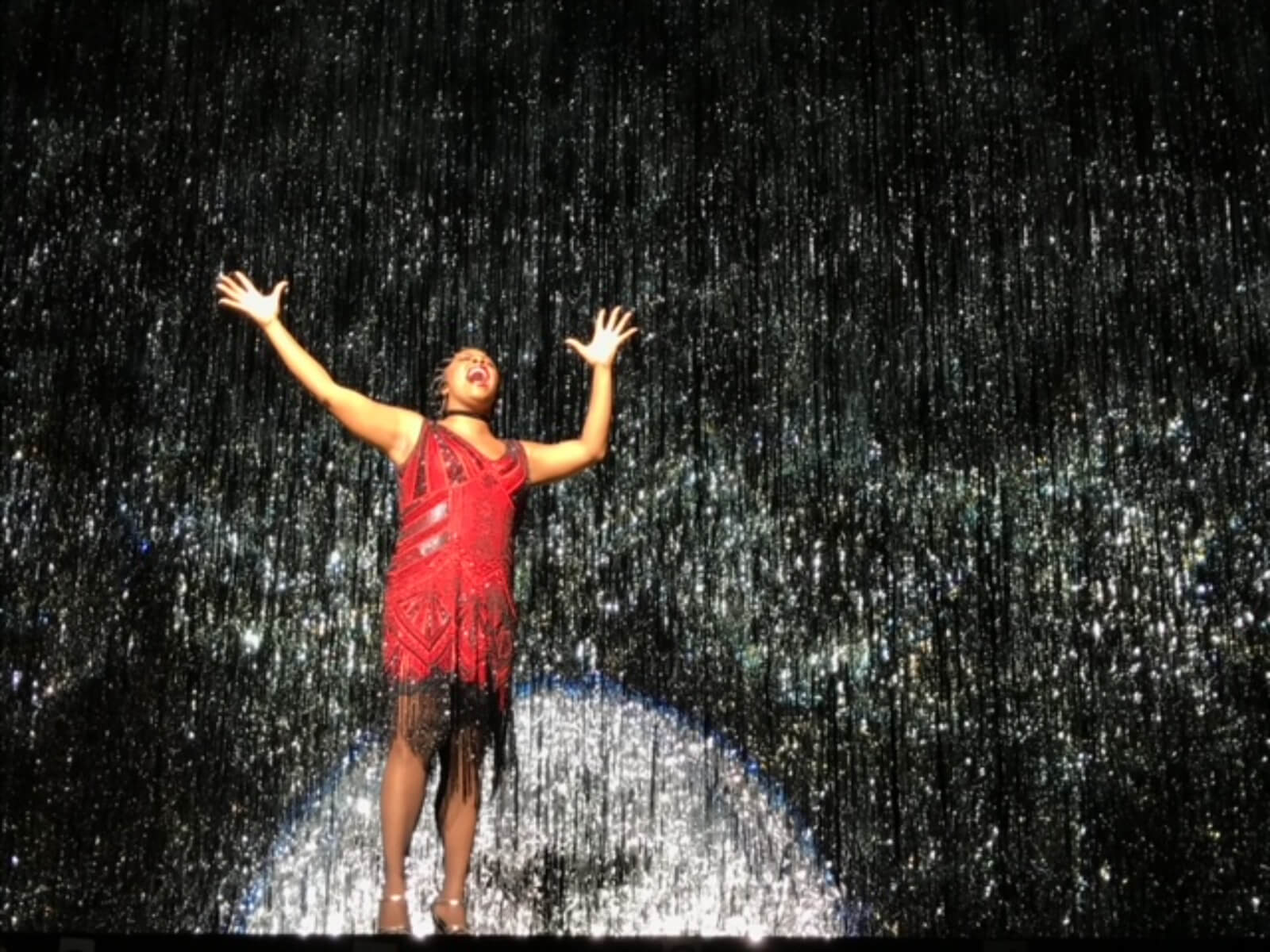 Bryonha Marie Parham singing "Cabaret" in a sparkly red dress in front of a shiny silver ribbon curtain in Prince of Broadway. 