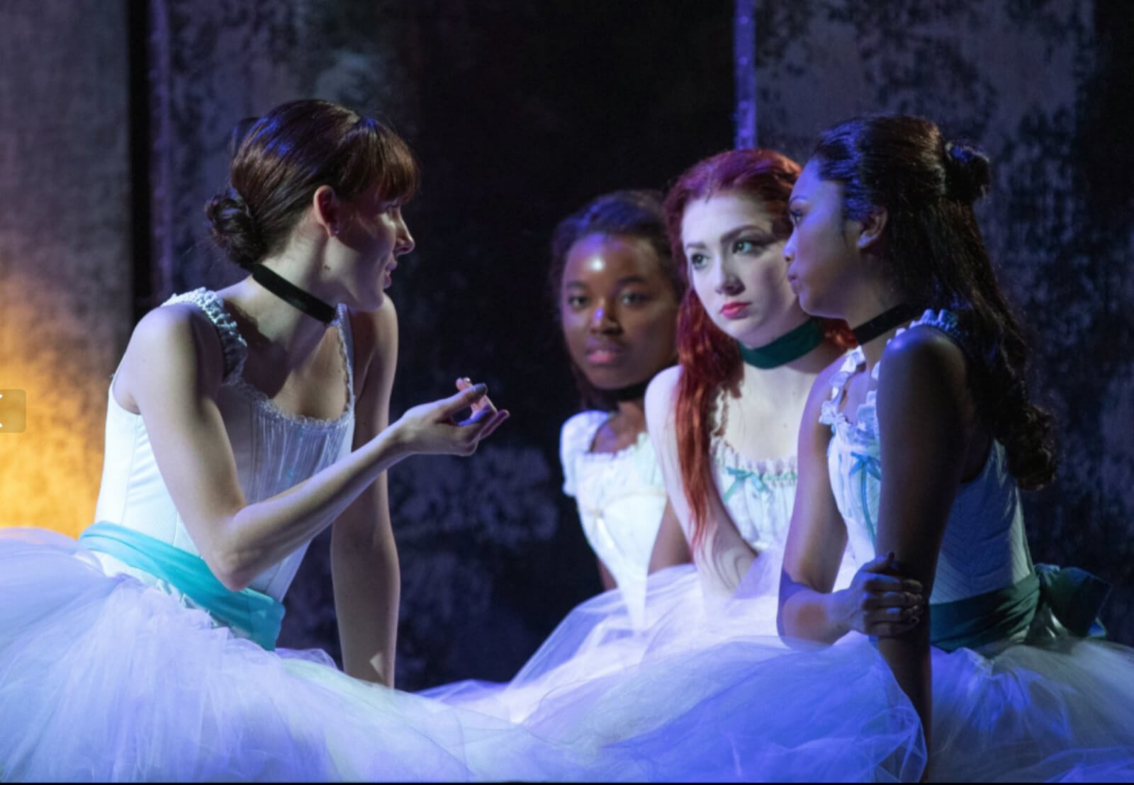 Marie and three girls in tutu and black neck collars, sitting. Marie is telling them something; she's explaining to the girls about the next ballet step.