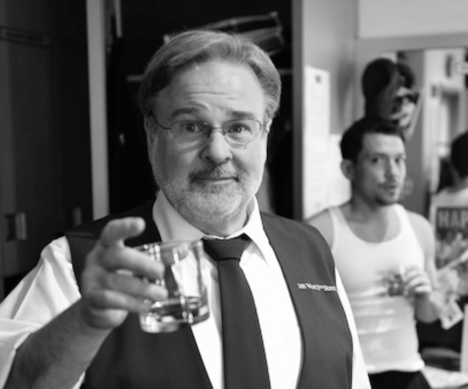 black and white photo of Fred Applegate toasting the camera with Miguel Cervantes in the background backstage.