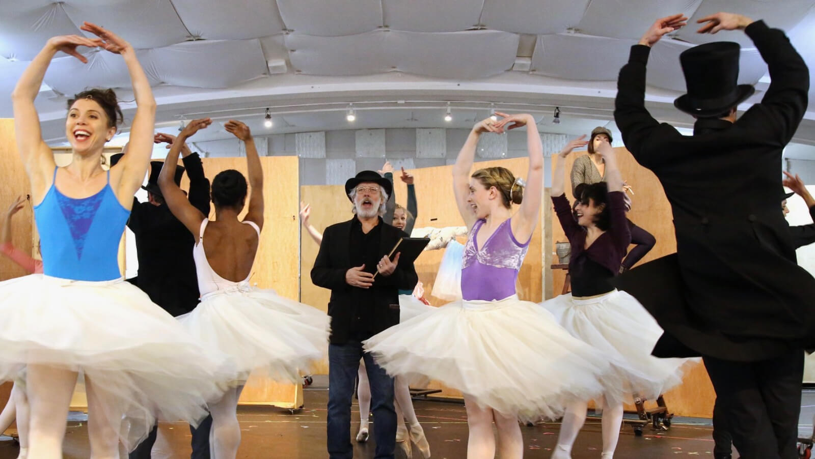 The cast in their beautiful and colorful leotard tops and white tutus, with male dancers in a black hat and a suit, during rehearsals, having fun while being serious with training at the same time. Degas stands in the middle.