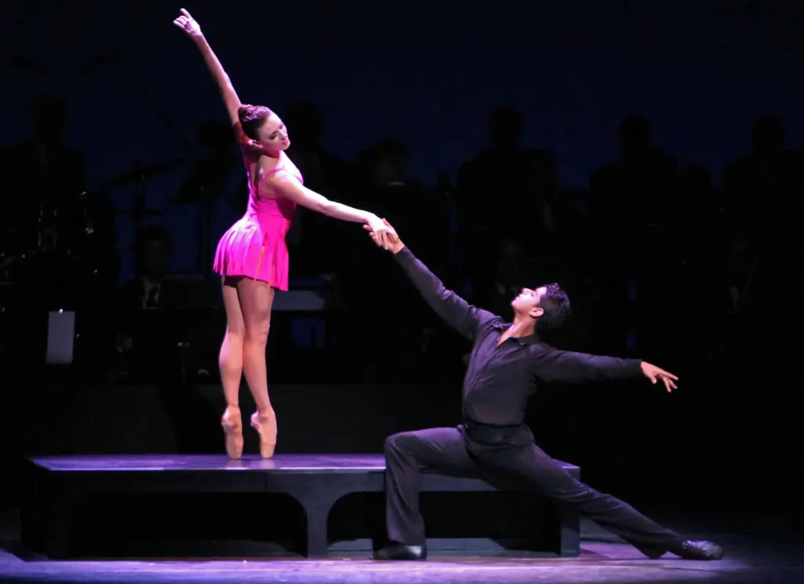 The pas de deux with Tiler Peck and Amar Ramasar continues on the bench: woman is standing on the bench en pointe while man is reaching in a lunge.