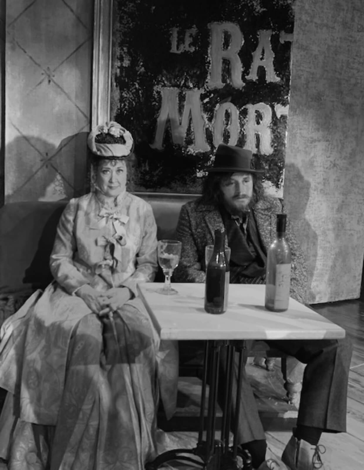 A black and white photo of Karen Ziemba and Jimmy Borstelmann in a bar. Karen is wearing a hat and a big dress, while Jimmy in a hat and a suit, is slouching and looks drunk. They are L'Absinthe drinkers.