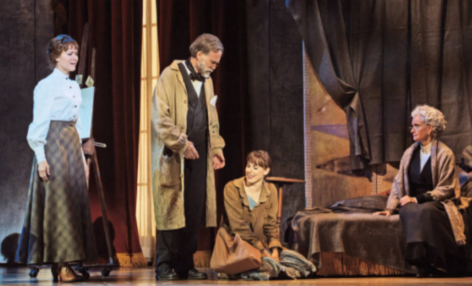 Marie wearing a bag and a coat, sitting on the floor, in conversation with three elders (two women and one man) inside a room with her.