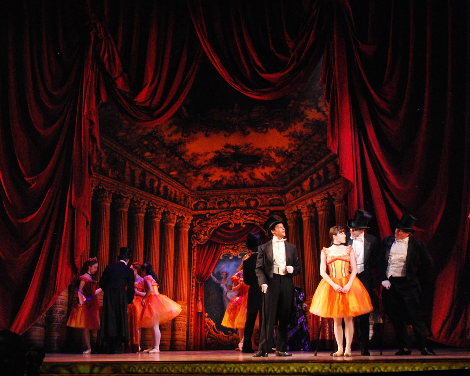 Marie (Tiler Peck) surrounded by the abonnés in the Foyer de la Danse. She is dressed in her butterfly tutu, the men are in their top and tails. There is red lighting on the set.