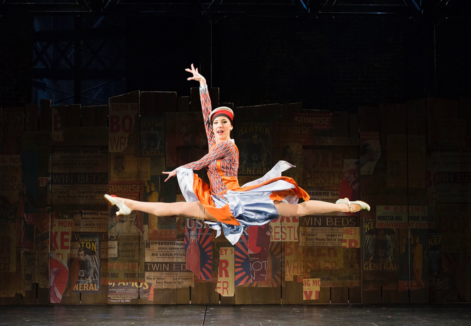 Brittany Marcin Maschmeyer dressed in a multi-colored dress does a split leap‍ in front of a signage covered backdrop.
