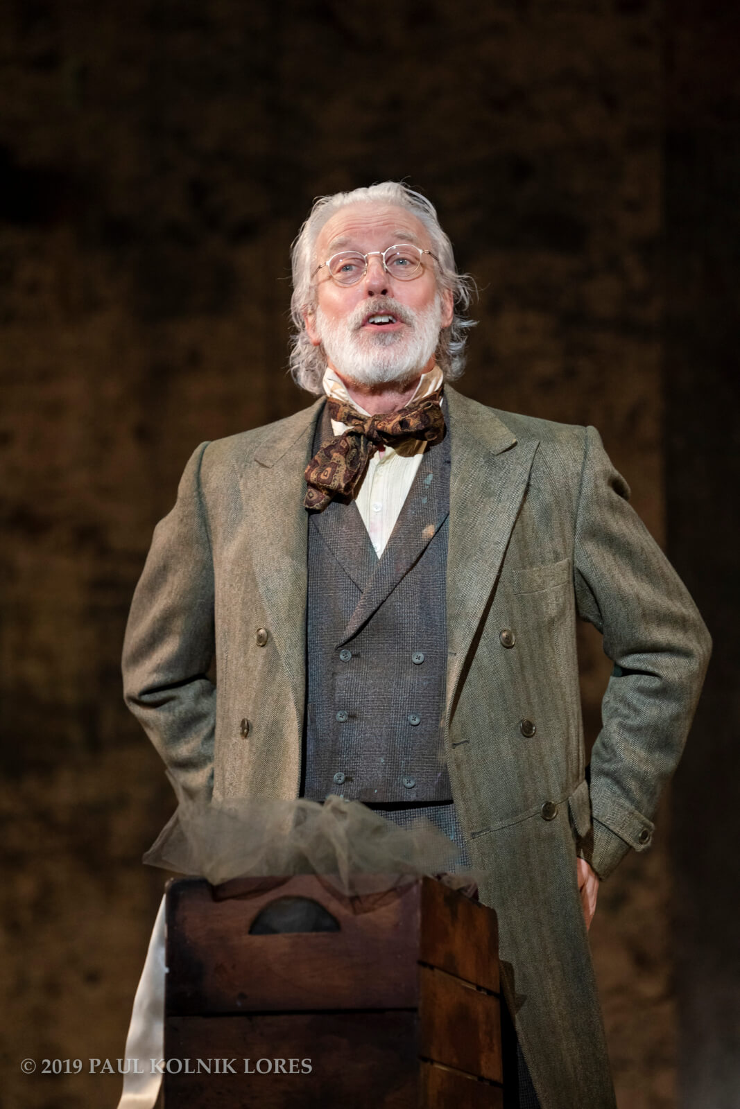 Degas in a vest suit, mossy green coat, and brown-patterned bowtie, wearing a pair of eyeglasses, is candidly speaking.