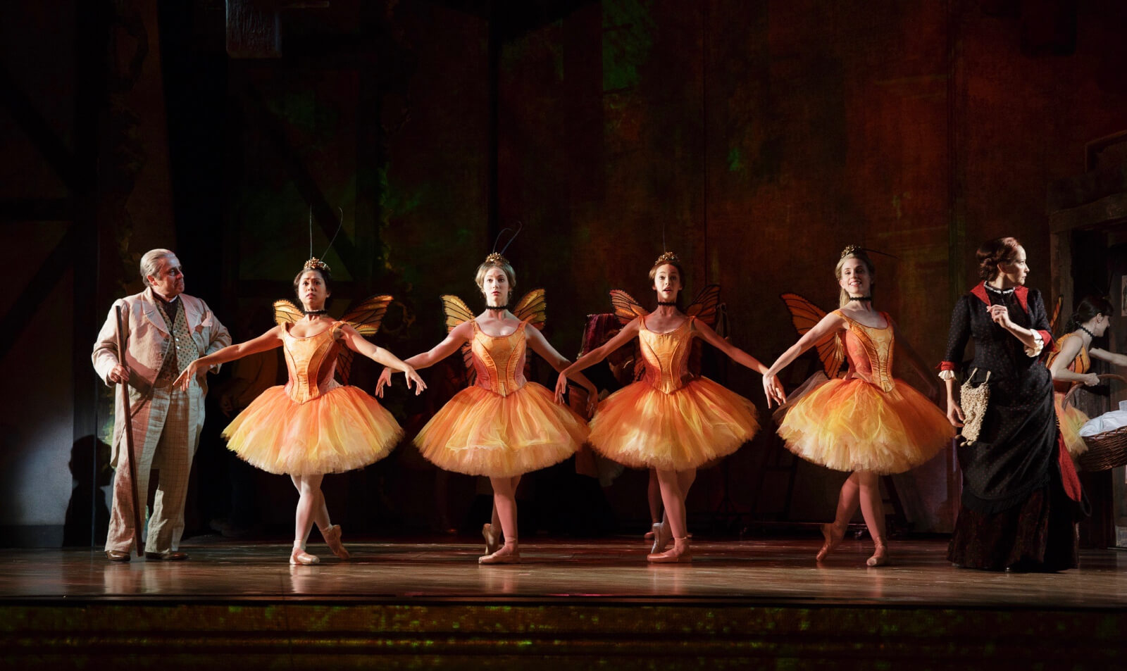 Four ballerinas in orange ballet outfits with matching wings and antenna, with arms in second position and feet in fourth position, being observed by their teacher.