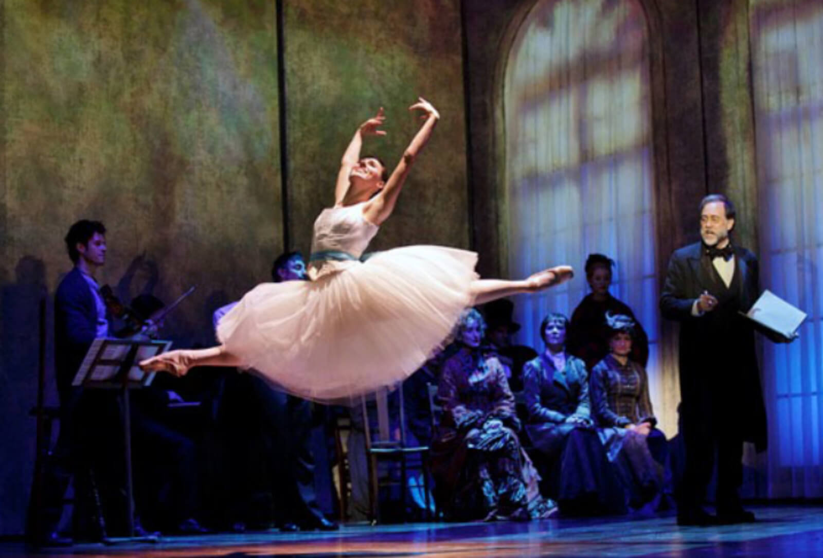 Marie in white tutus and turquoise ribbon as belt, doing a perfect grande jeté with arms in 5th position, inside low-lit studio with a small audience. Her mentor looks pleased.