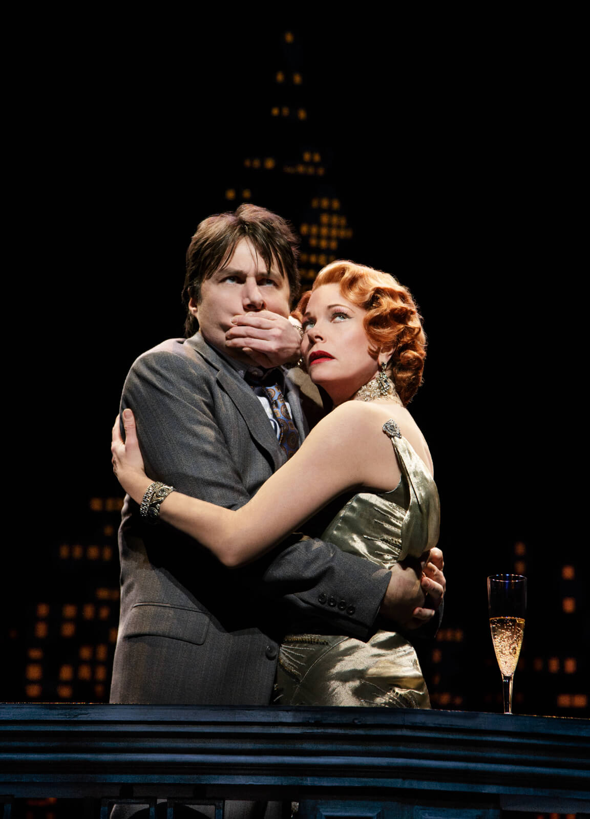 Helen Sinclair (Marin Mazzie in a cocktail dress) hugs and covers the mouth of David Shayne (Zach Braff) with a cityscape in the background. Saying the iconic line "Don't Speak".