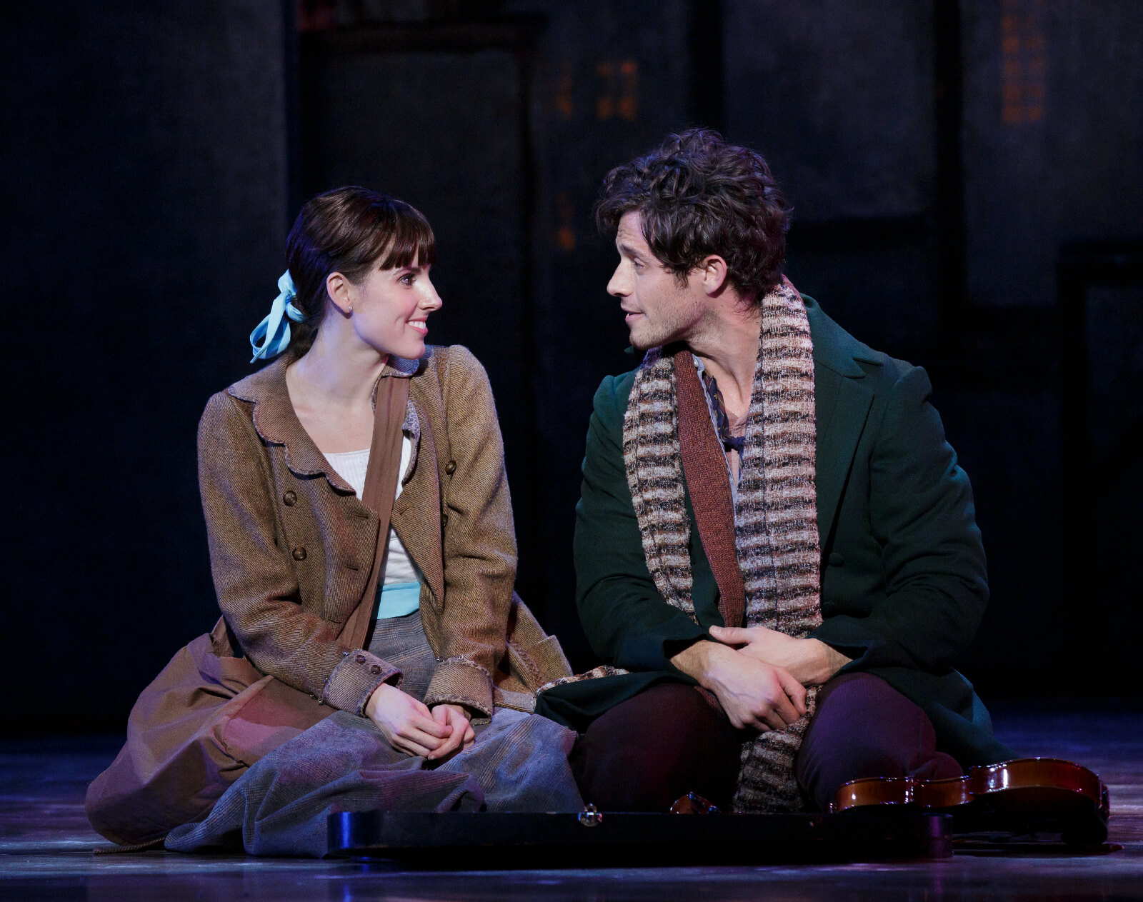 Marie and Christian with their bags and in their casual clothes, sitting on the floor, looking at each other's eyes. Christian's violin is right in front of him.