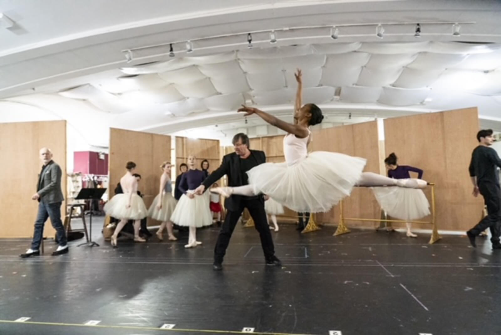 Jasmine Ward jetés across the room during rehearsals. One ballerina is in focus, doing a jeté as her teacher instructs a few inches away from her.