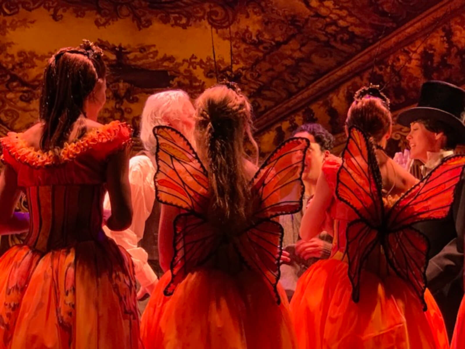 Ballerinas in butterfly costumes, with matching antennas and wings, being instructed before the show. They are in a huge hall with fancy painted walls and ceilings.