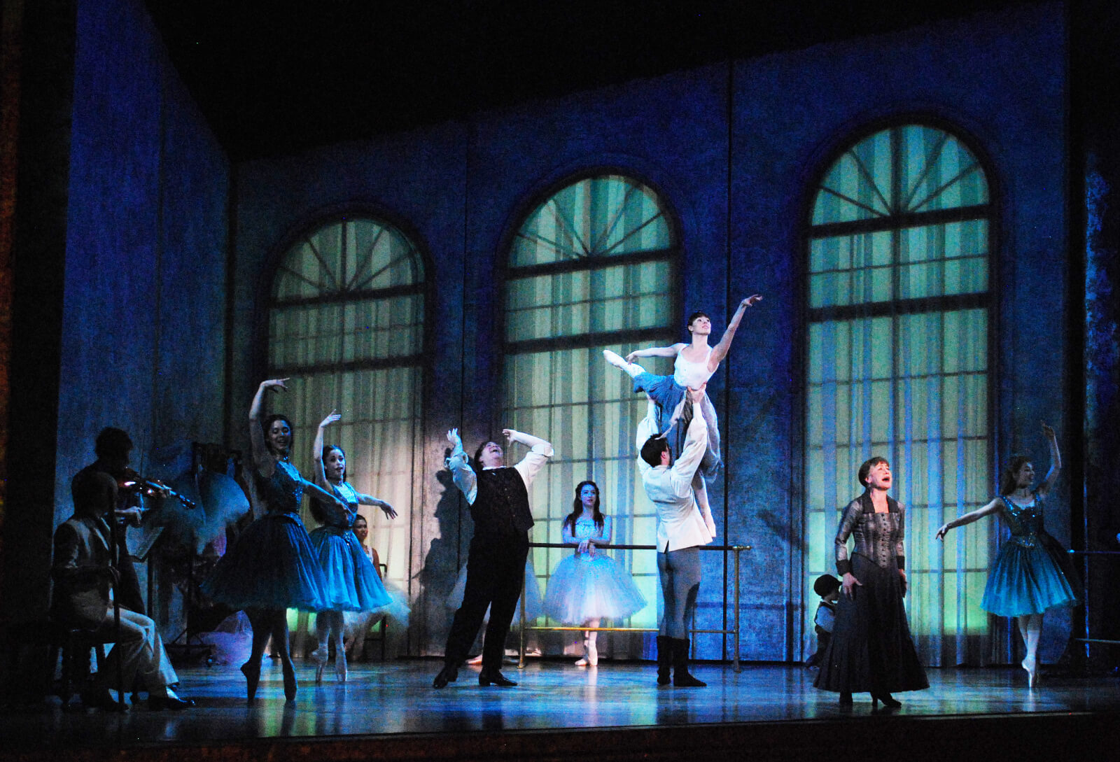 In a low-lit room, Marie is being carried by another dancer during rehearsal, while two ARTISTS are singing and another musician at the back is playing the violin.