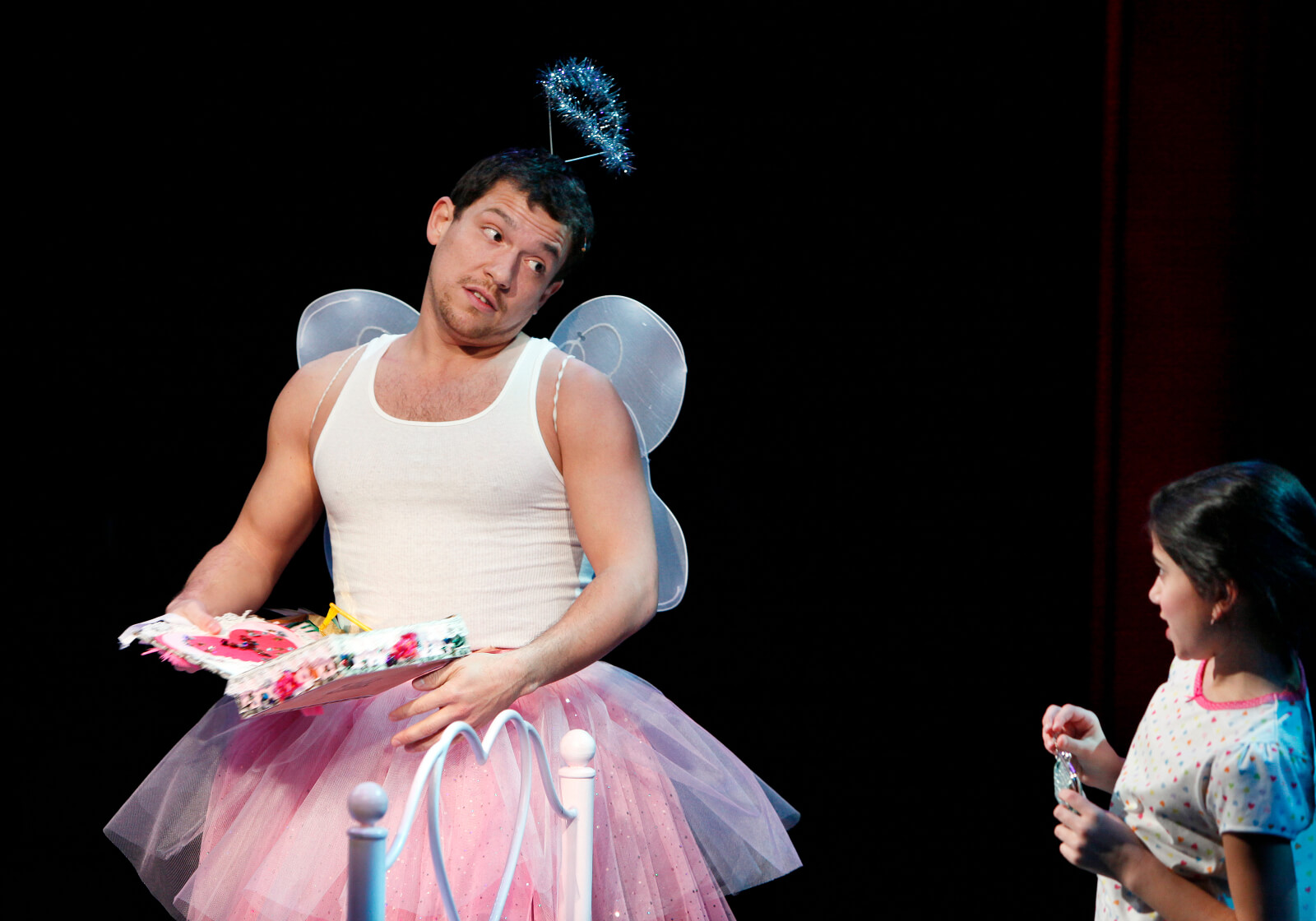 Miguel (Miguel Cervantes) in a tutu and tank top with a sparkly crown reads to a young girl, Yolanda (Lina Silver) from a colorful book.