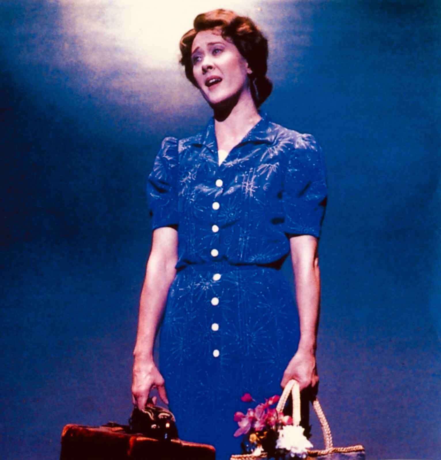 Karen Ziemba as Lizzie singing "Simple Little Things". She wears a button down blue dress with puffy sleeves, holding luggage and a basket with flowers.