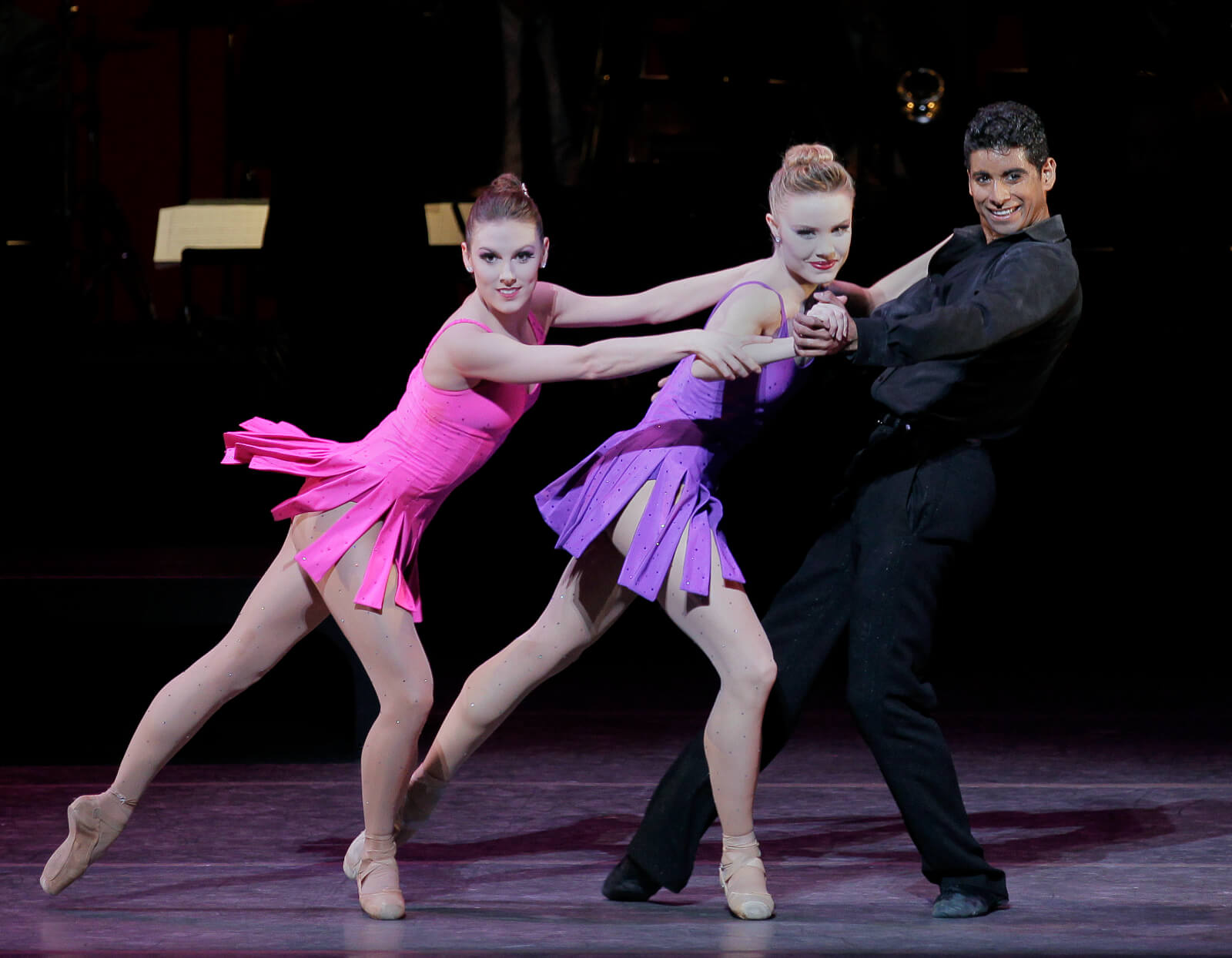 World premiere of For the Love of Duke: two women, Tiler Peck in pink and Sara Mearns in purple, dance with Amar Ramasar, dressed in all black.