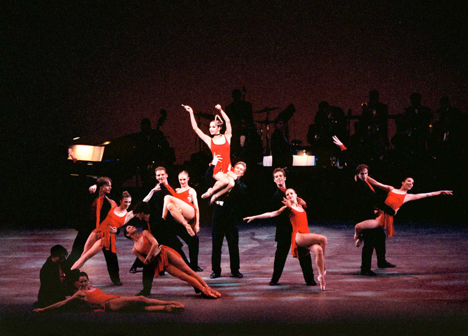 The full company of Blossom Got Kissed in various lifts and partnering, all dressed in red and black. One woman is on a man's shoulder and ready to ring a triangle.