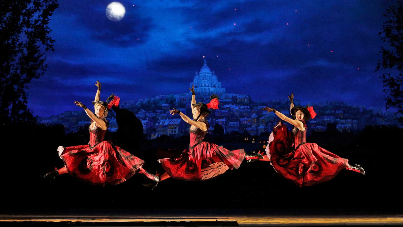 Erin Monteleone, Emery LeCrone and Kristen Schoen-Rene doing a jete in the air wearing their Red and Black Can Can dresses. There is a view of Paris at night behind them. 