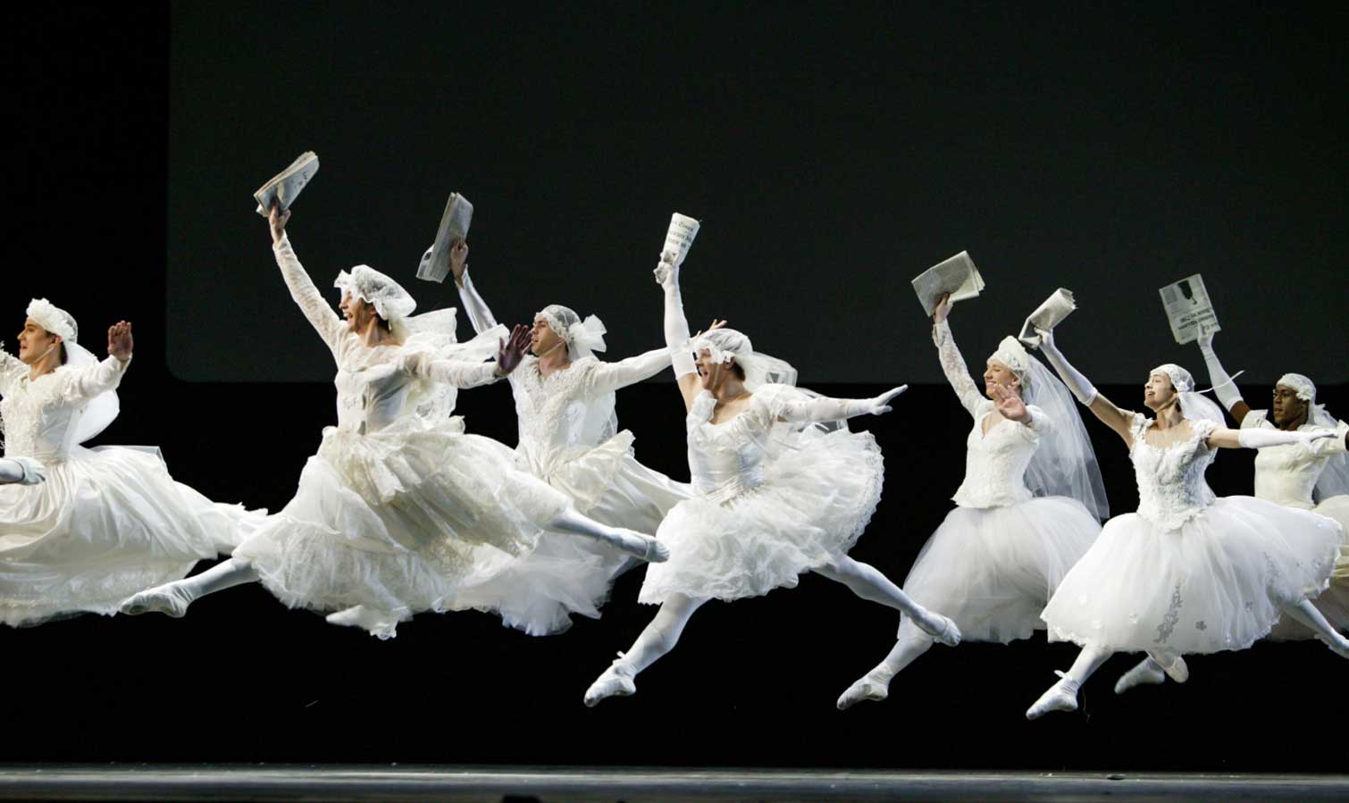 A group of brides all in white (some men dressed as women) leap at the news, held in their upstage hand.