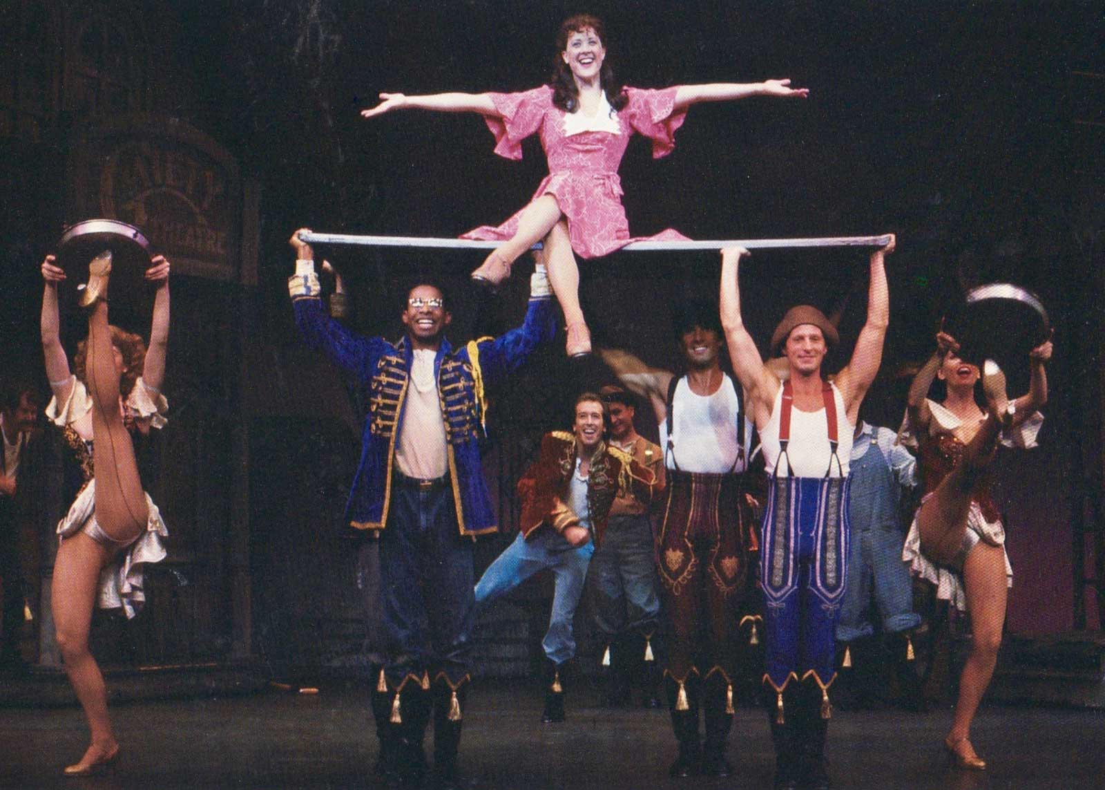 Polly Baker (Karen Ziemba) opens her arms out wide and sits on a plank that is lifted by three men in the musical number "I Got Rhythm"
