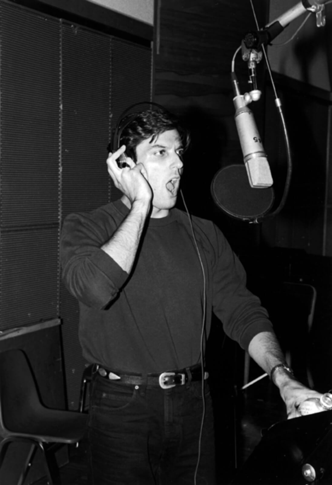 Black and white photo of Bob Cuccioli recording the cast album to And the World Goes ‘Round. He is wearing a turtleneck and has one hand on headphones while singing into a microphone.