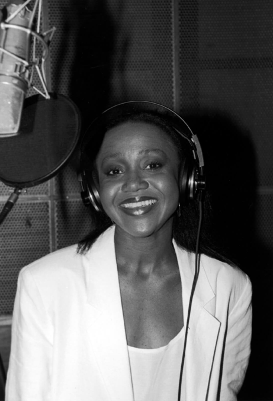 Black and white photo of Brenda Pressley recording the cast album to And the World Goes ‘Round. She is dressed in all white and standing in front of a microphone.