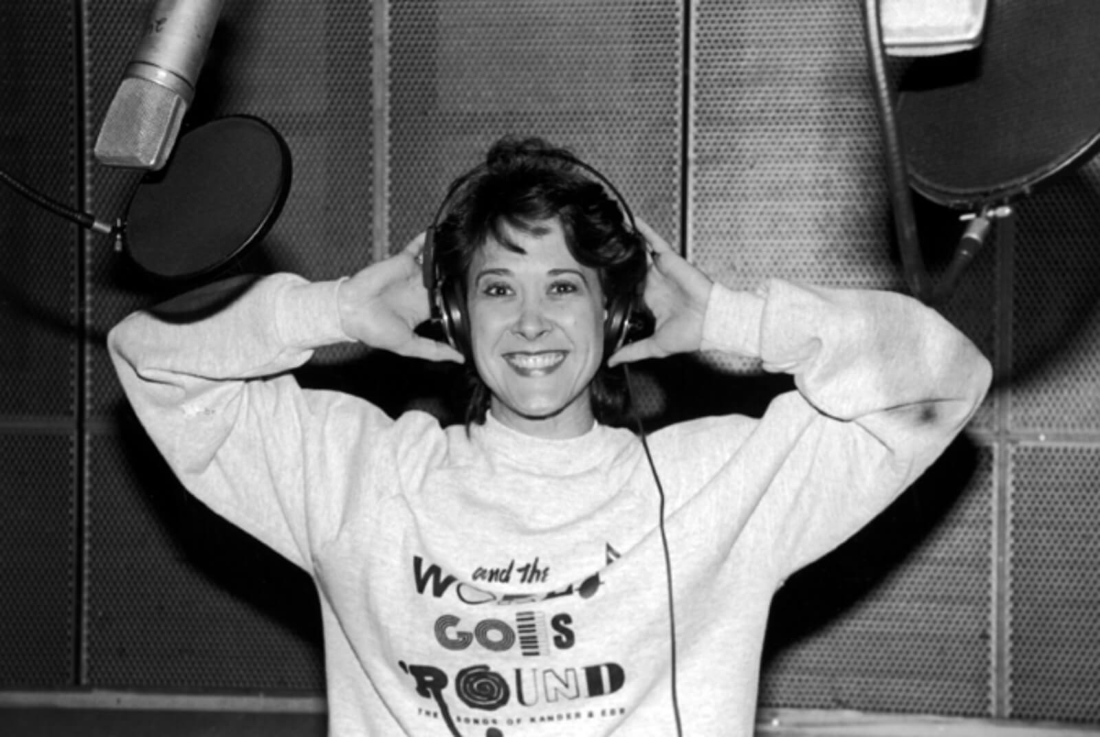 Black and white photo of Karen Ziemba recording the cast album to And the World Goes ‘Round. She is wearing the show's logo on a sweatshirt and holding headphones.