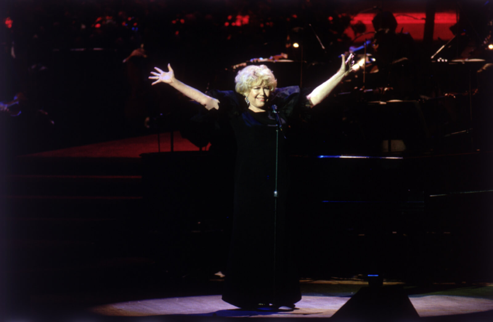 Dorothy Loudon stands singing with both arms raised and open in a wide "V"