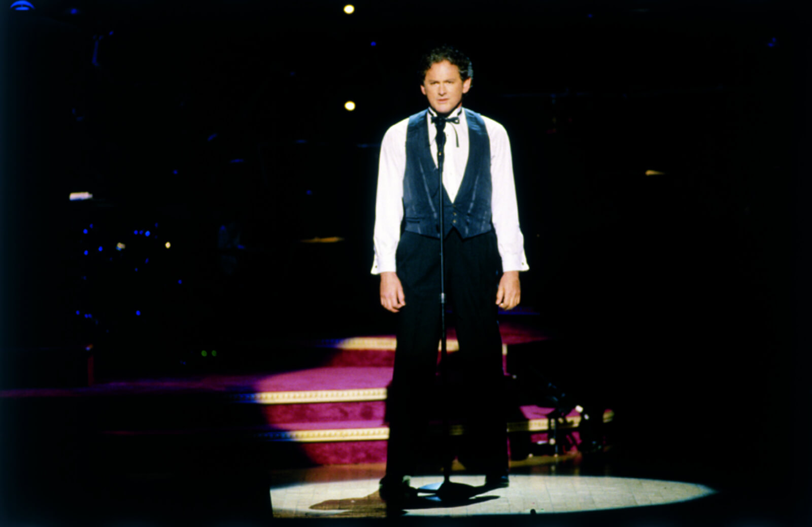 Victor Garber in a blue vest and tie, stands, singing at a microphone.