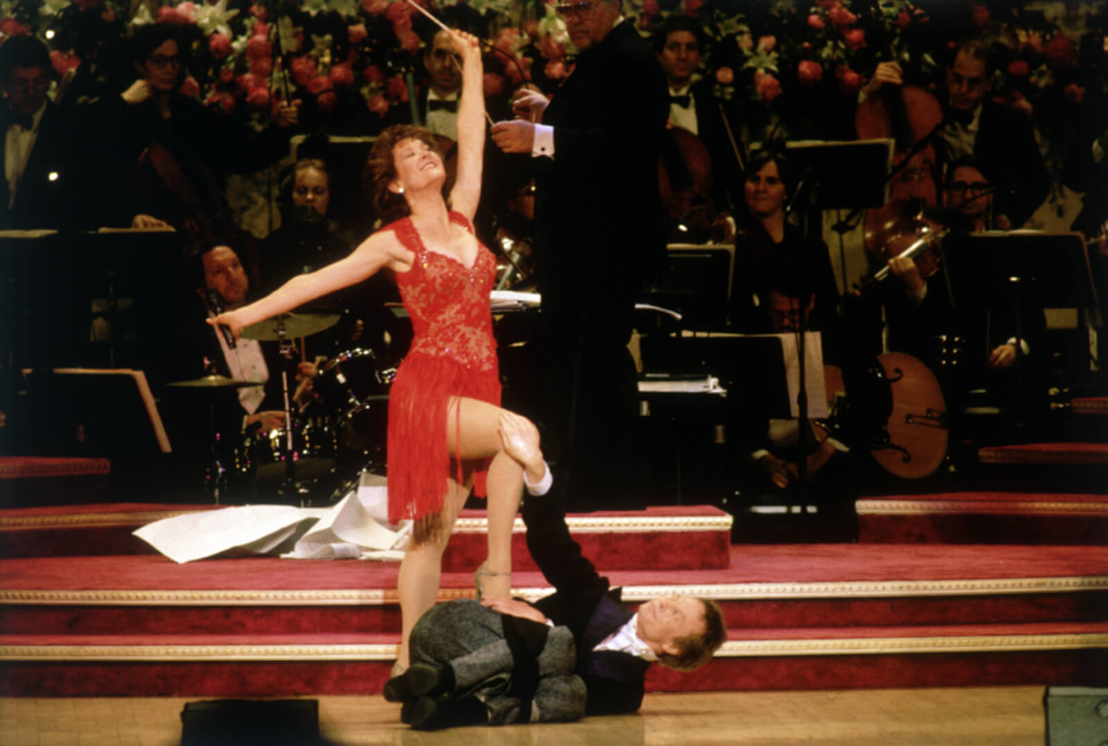 Karen Ziemba and Bill Irwin performing "Sooner or Later". She in standing over him in a red dress with one leg on him. He is laying in a cradle post on the ground in a suit.