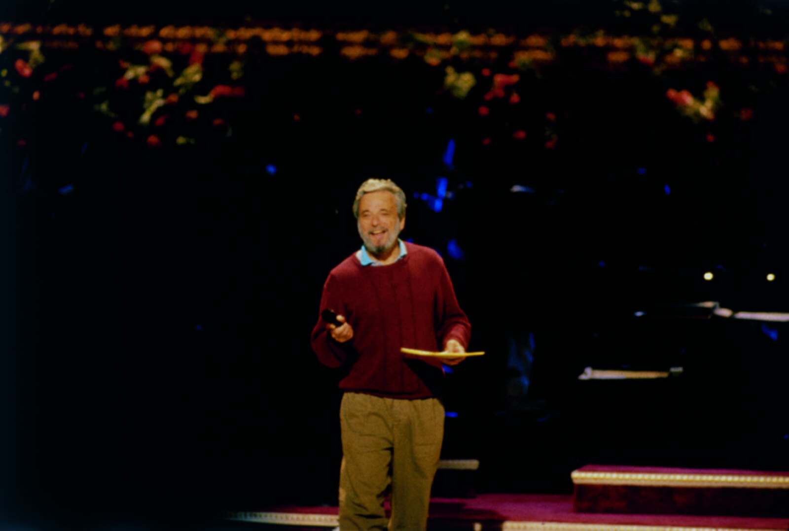 Stephen Sondheim in rehearsal. He is wearing a blue dress shirt and a maroon sweater, holding a microphone and notepad.