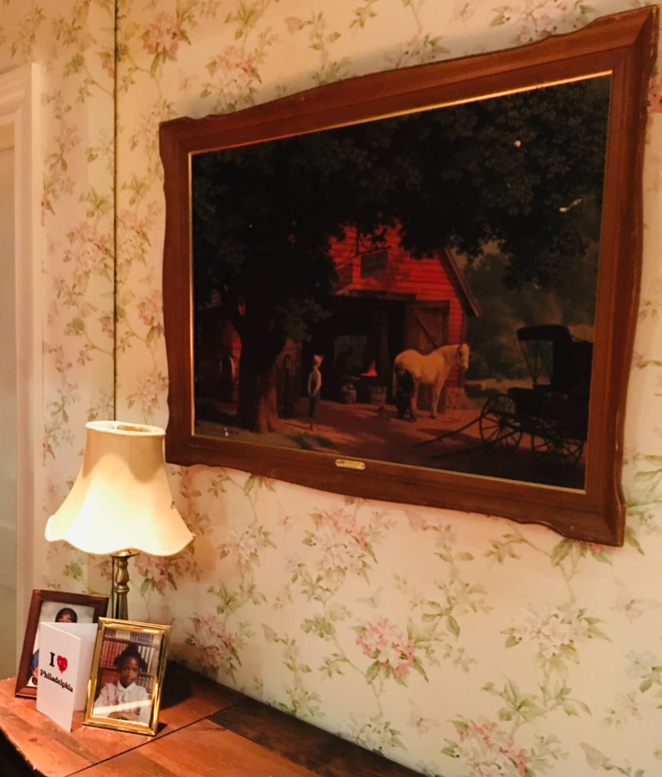 Dot set by Allen Moyer. Photo shows a framed painting of a horse and barn against a floral wallpaper. A desk below the photo shows a table light, framed photo of a young black boy, framed photo of a young black girl, and a greeting card with "I Love Philadelphia" on the cover. 