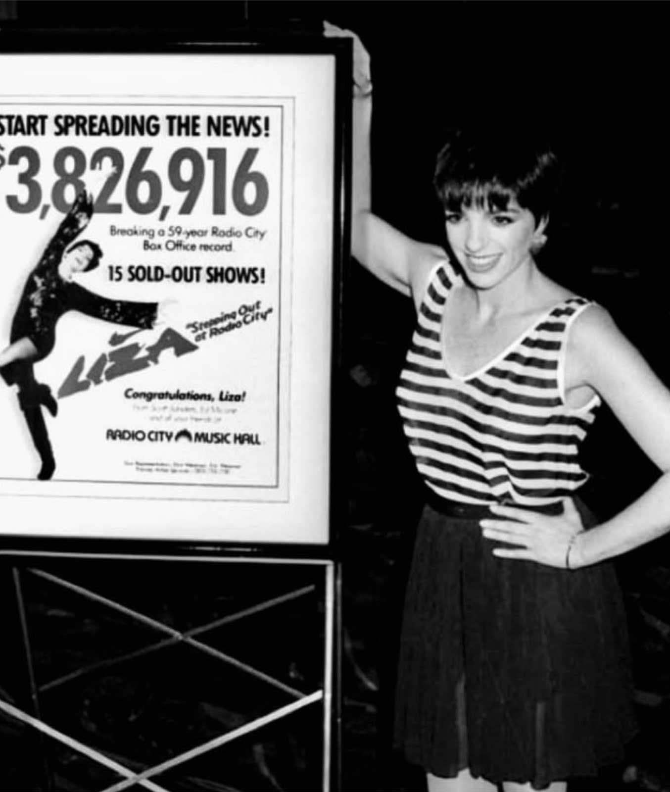 Liza establishes new Radio City box office records with 15 sold-out shows. She stands in striped shirt next to poster announcing the news, including a headline, "Start Spreading The News! $3,826,916"