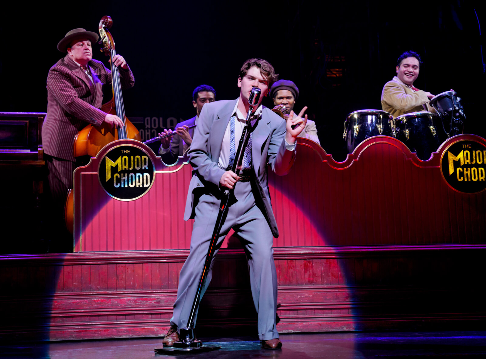 Colton Ryan performing “Music, Money, Love.” He is singing in a grey suited with an old school microphone while a band plays upstage of him with "The Major Chord" on the bandstand.