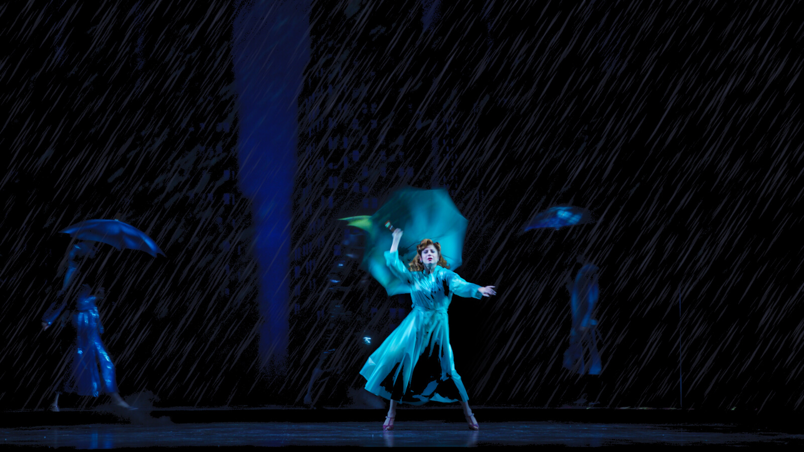 Ashley Blair Fitzgerald in “New York in the Rain.” She is seen downstage in a blue raincoat and umbrella - dancing in the rain - with dancers walking past in the background.