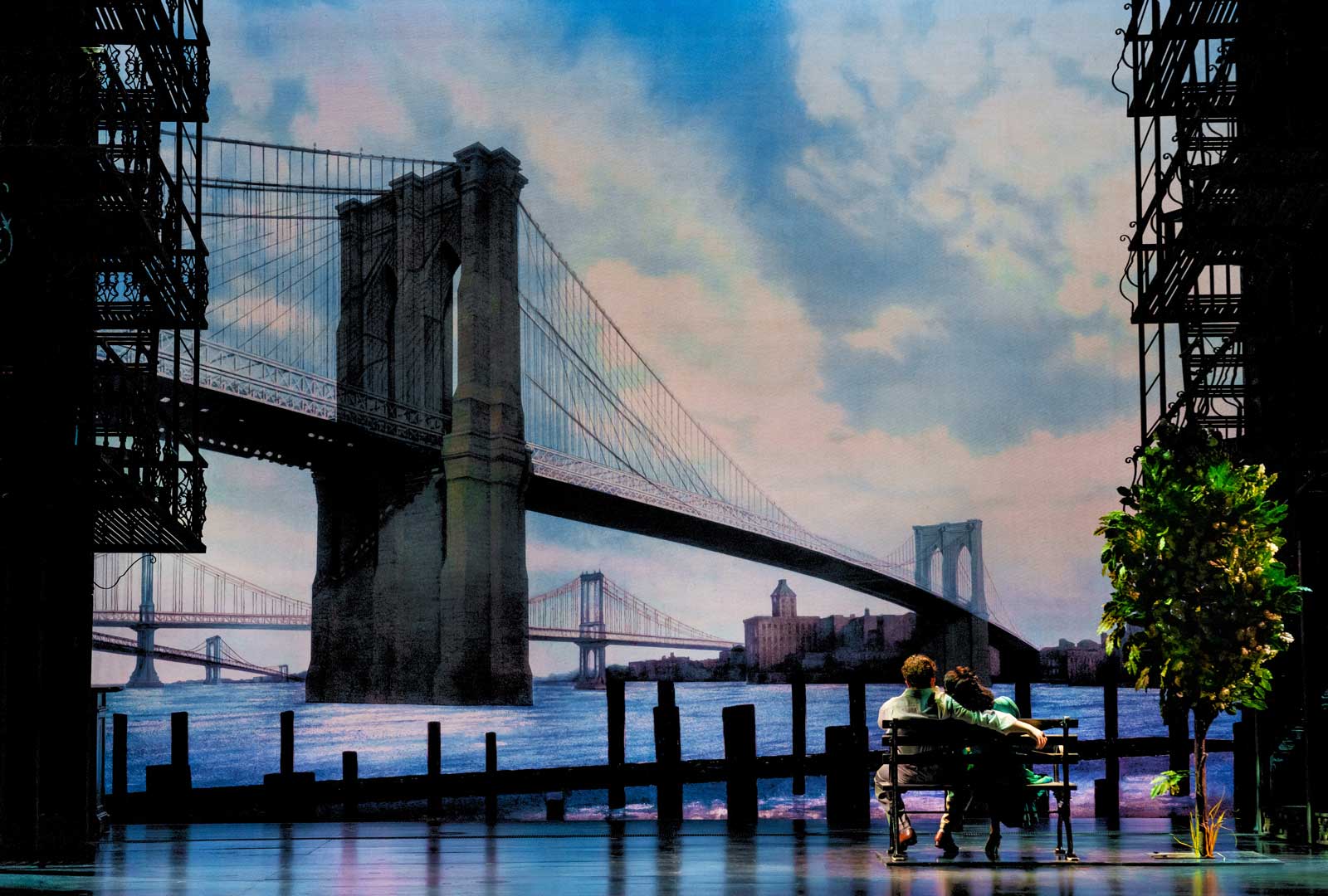 “Along Comes Love” with Colton Ryan and Anna Uzele. The lovers are on a park bench, seated facing upstage. They are looking at the Brooklyn Bridge while seated closely together.