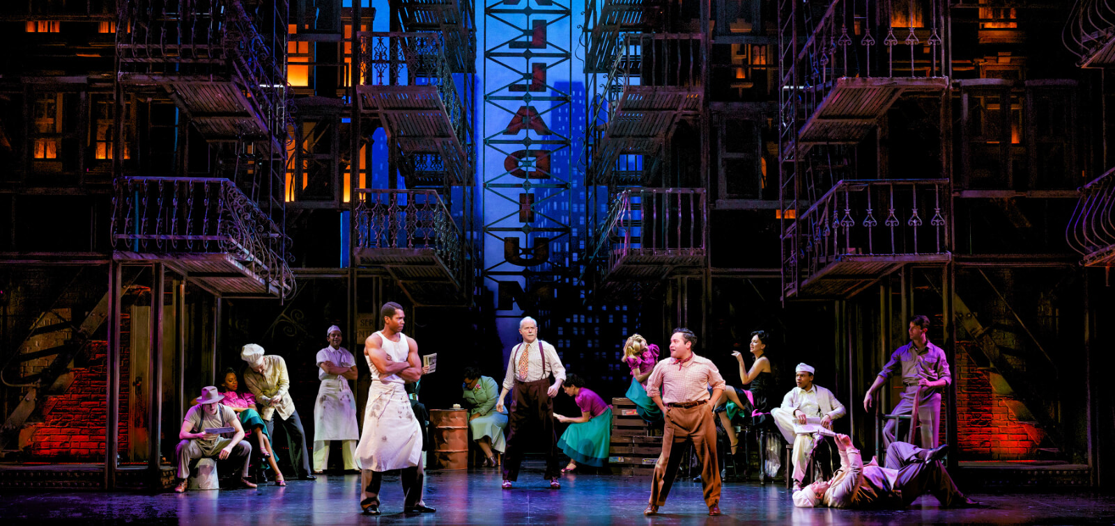 The company performing in “My Own Music.” A diverse cast of New Yorkers are somewhat paying attention to a discussion happening downstage. Fire escapes and a neon sign saying "Palladium" are in the background.