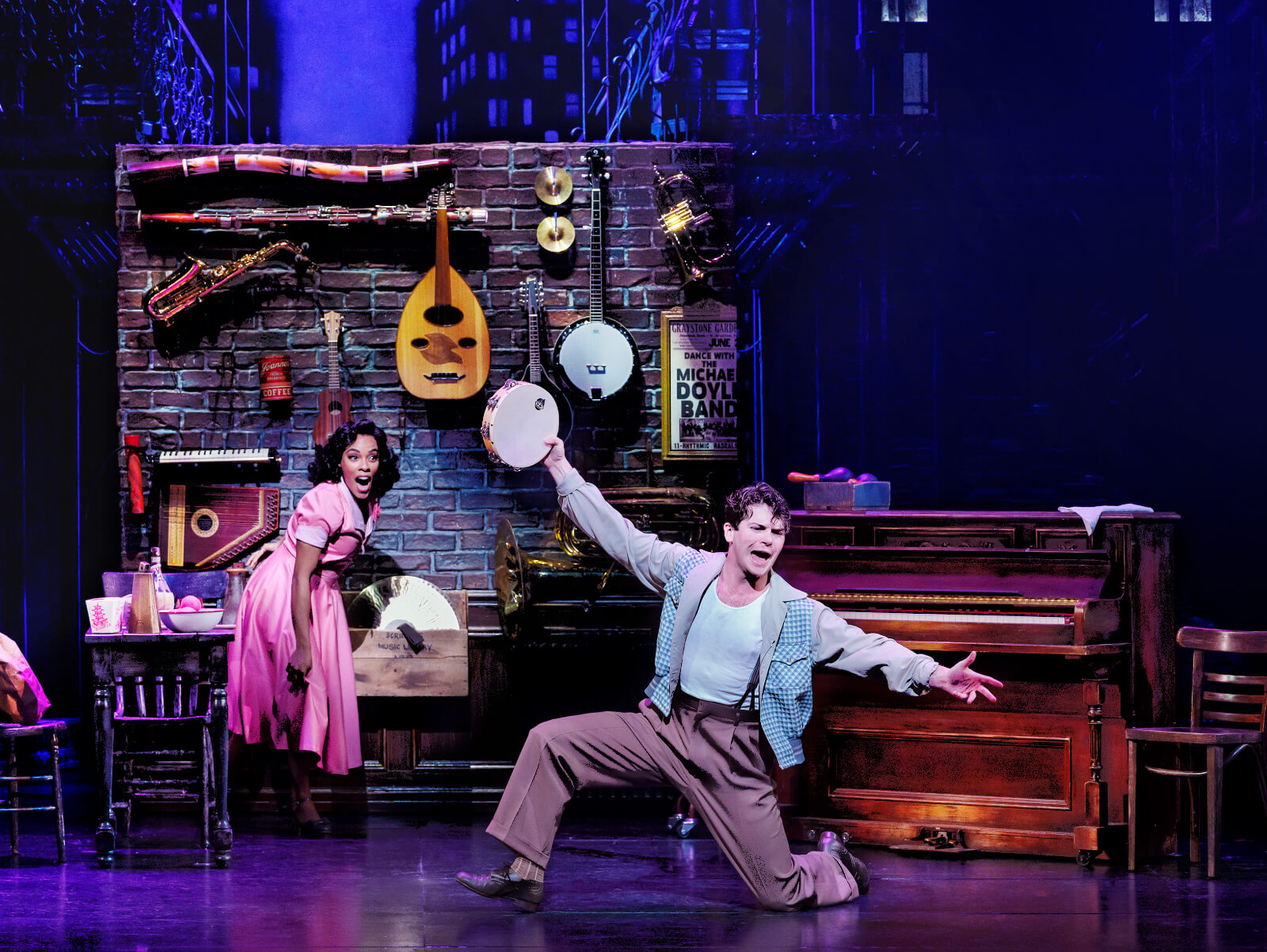 “I Love Music” with Anna Uzele and Colton Ryan. Colton is downstage in an open lunge playing the tambourine while Anna is upstage with her jaw dropped. There are musical instruments all along a brick wall.