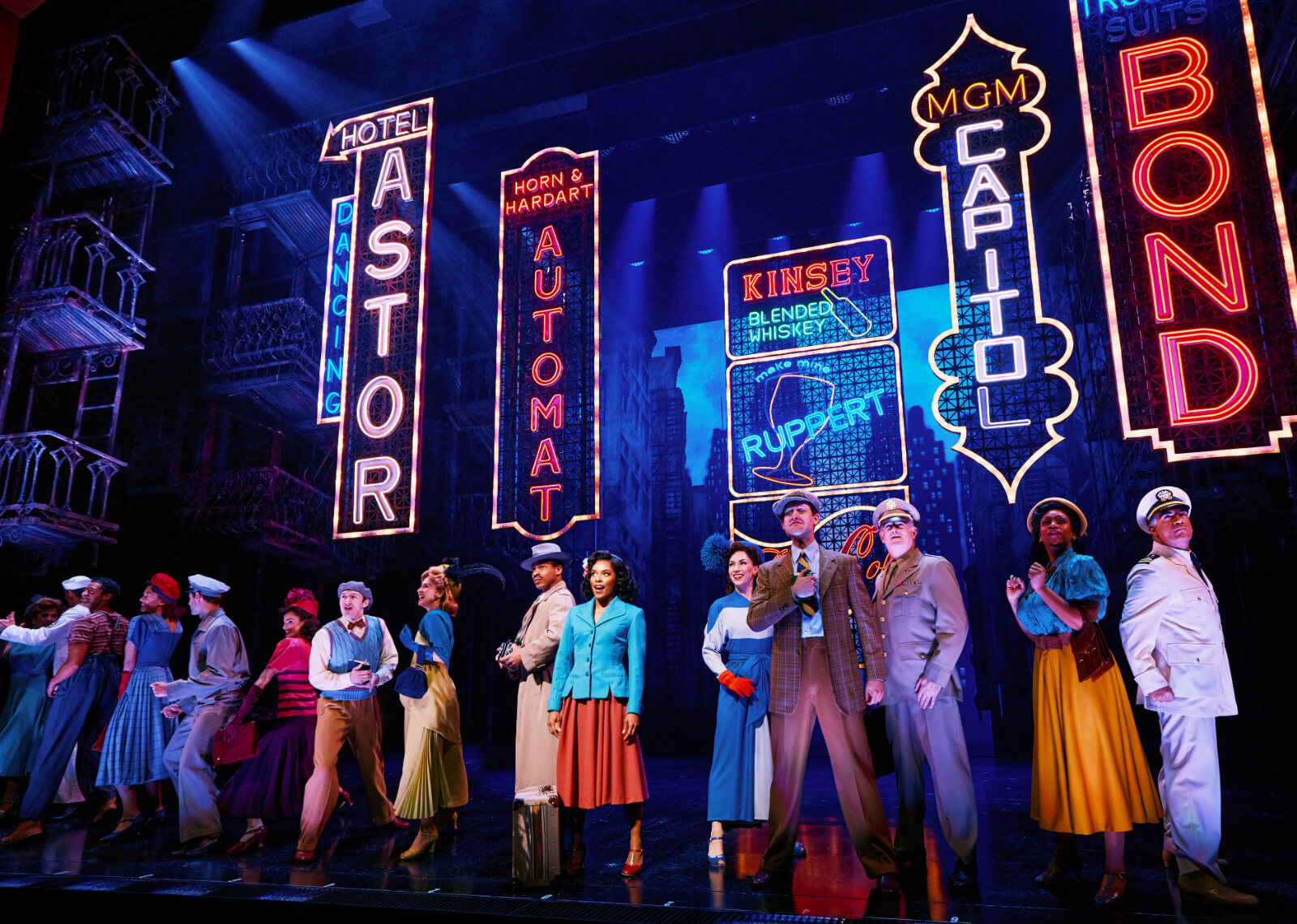 Opening number “Cheering For Me Now” with the cast of New York, New York. A diverse cast is in a staggered line downstage with the lights of Times Square behind them.