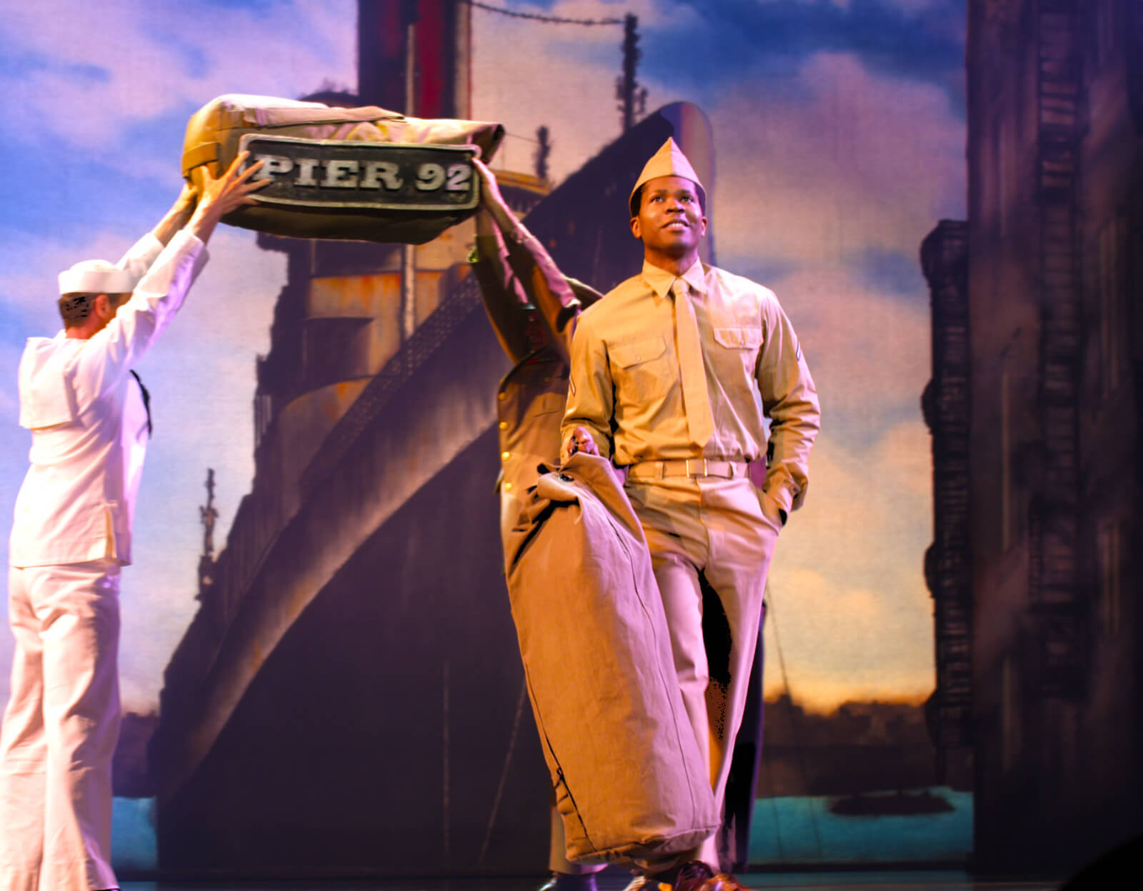 John Clay III in “Cheering For Me Now.” Dressed in a navy uniform, he walks downstage while a sailor and fellow service man create a bridge saying "Pier 92" out of a backpack.