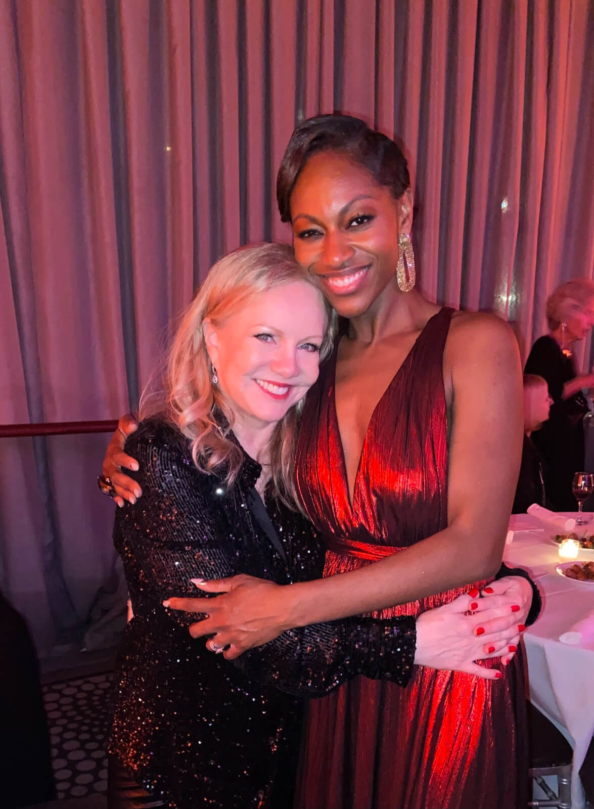Opening Night of New York, New York with Associate Director, Jenny Laroche. They are hugging. Stro is on the left in black sequins and Jenny is on the right in a red satin dress.