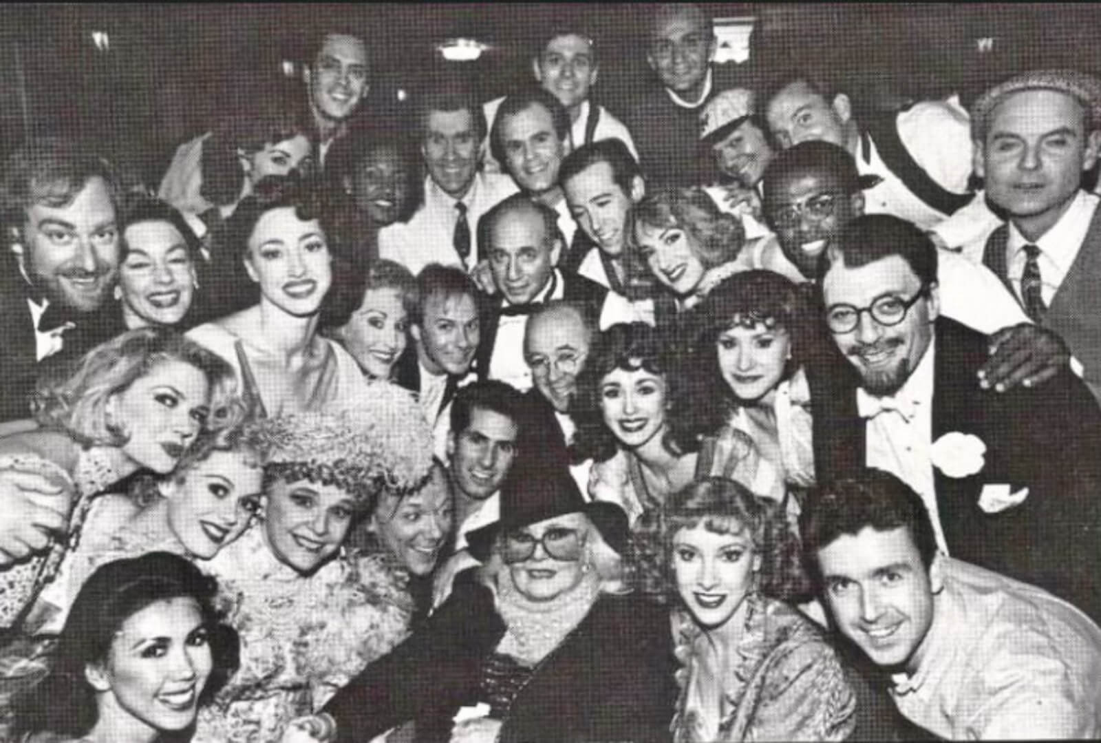 Black and white photo of the Original Broadway Cast of Crazy For You getting a visit backstage from Ginger Rogers. She is seated in front with a large mass a people surrounding her for the photo.