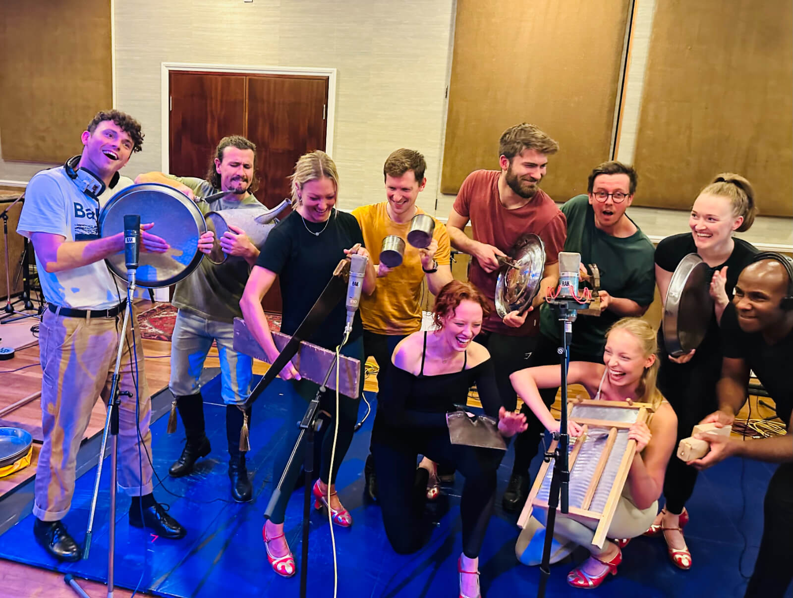 Photo of Charlie Stemp, Nicholas Duncan, Laura Hills, Liam Wrate, Ella Valentine, Lucas Koch, George Bray, Tara Yasmin, Nell Martin, Resident Director-Choreographer Richard Pitt at RAK Recording Studios in St John’s Wood. They are holding musical instruments with microphones and recording equipment surrounding them.