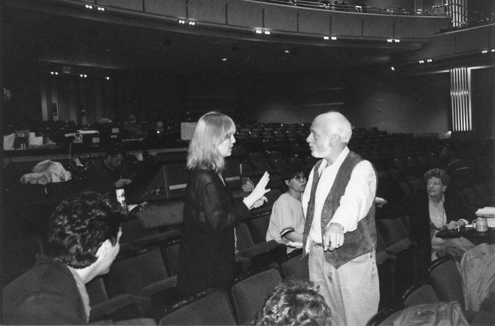 Black and white photo of Susan Stroman and Hal Prince having a spirited conversation in tech rehearsals. The empty Broadway house seats are seen with tech tables and computers/lights in the background. Behind the two standing in conversation are future Broadway director Stafford Arima and Stage Manager/Assistant Ruth Mitchell seated in the background.