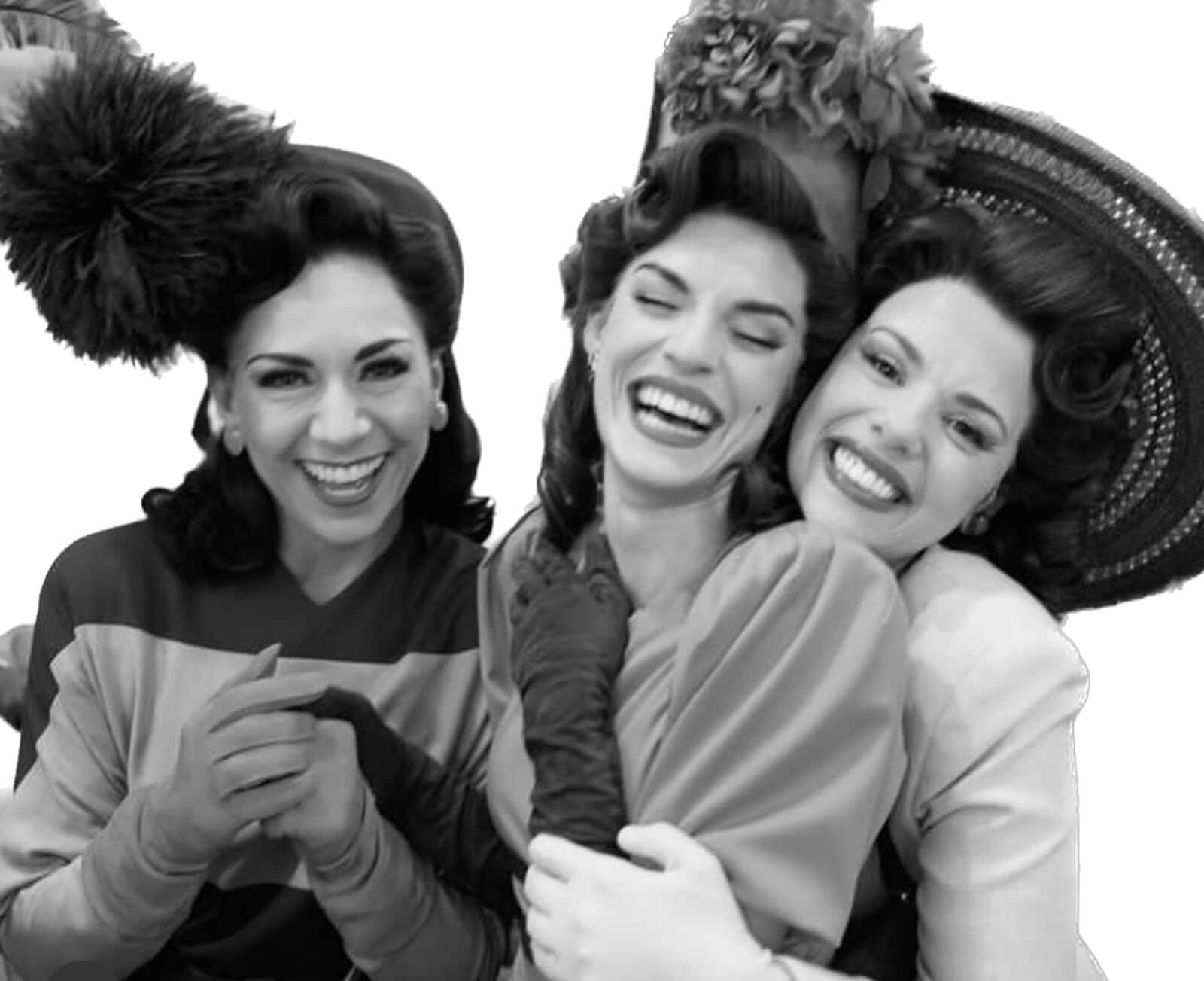 Black and white photo of cast members Wendy Bergamini, Gabriel Enriquez, Janet Decal backstage at New York, New York. They are wearing 50s attire, including big feathered hats. They are hugging and smiling.