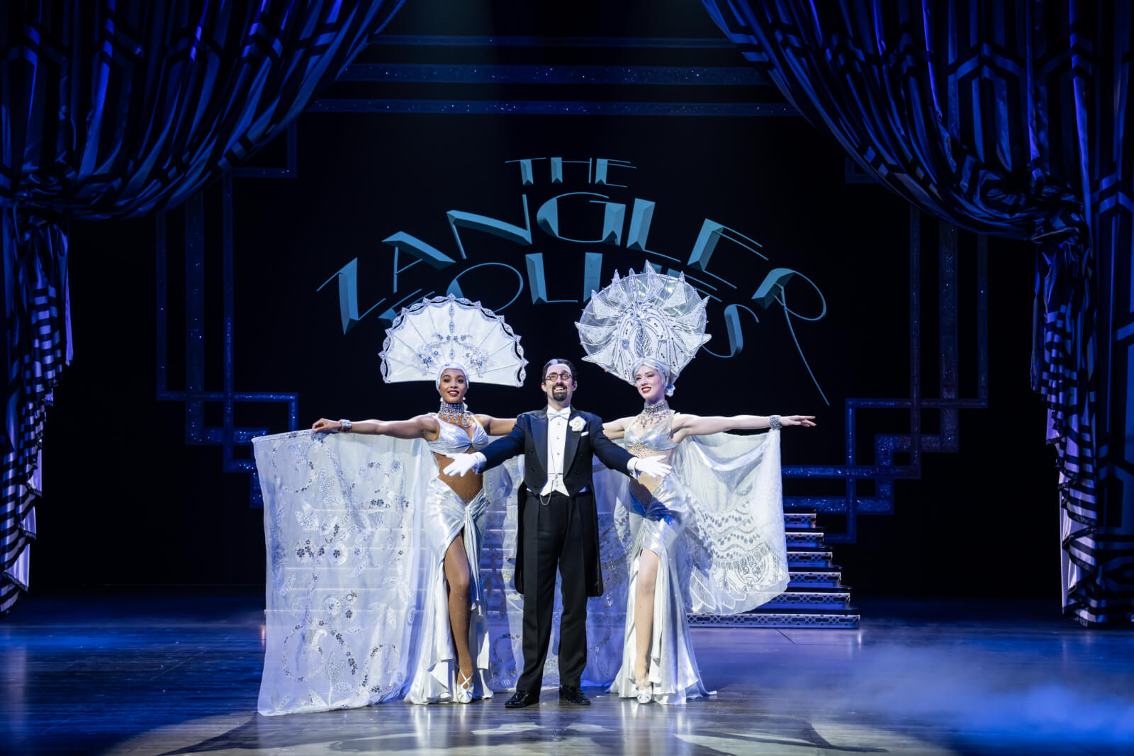 Tom Edden (Bella Zangler) with Harriet Samuel-Gray (left) and Ella Valentine (right). Bella is dressed in a tux with tails while the women surround him in silver showgirl costumes, complete with open capes and large headdresses.